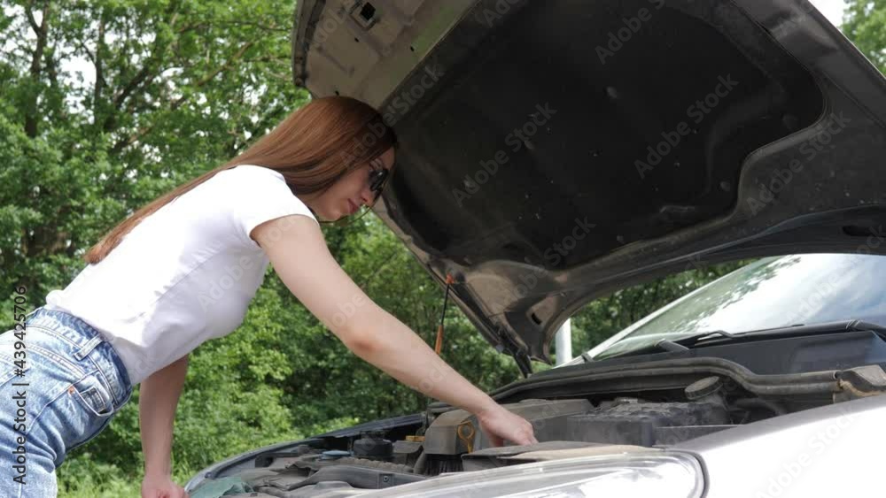 A young beautiful girl stands by a wrecked car on the highway. A girl with glasses looks under the hood of a broken car, trying to fix it. Problems with the car. A girl on the road repairs a car