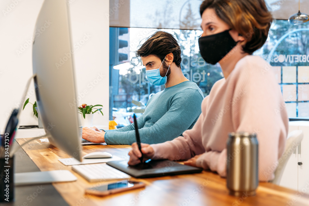 Two cheerful colleagues with mask working on computer in an office Two cheerful colleagues with mask working on computer in an office