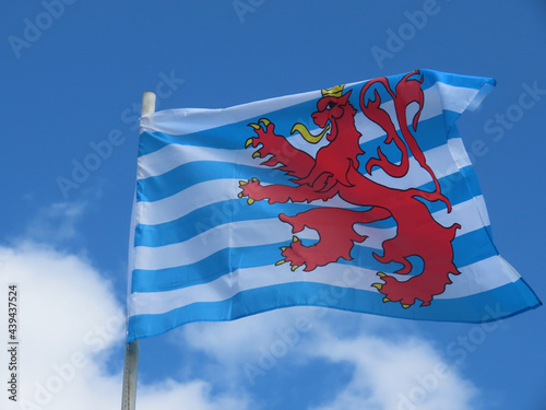 The Red Lion - civil flag and ensign of Luxembourg featuring white and blue stripes, red lion with a yellow tongue, claws and crown. 'Roude Leiw' flying in wind on a bright summer day. Selective focus photo