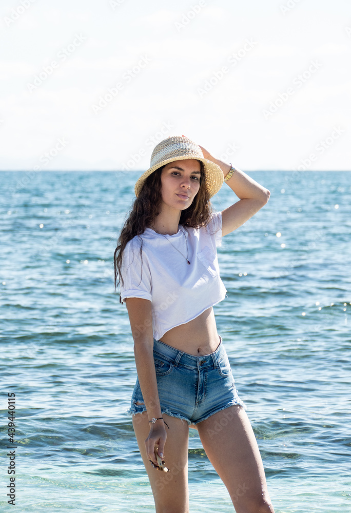 Attractive latin woman portrait outdoors standing by the sea wearing summer clothes and a hat