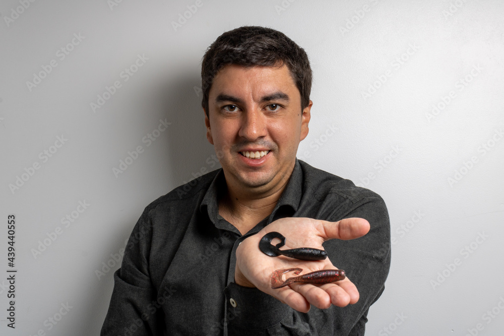 Brunette man holding two soft lures that mimic an earthworm or shrimp, commonly used with a jig head. Mention the preparation for a day of family fishing on the beach, river or lake. Brunette man holding two soft lures that mimic an earthworm or shrimp, commonly used with a jig head. Mention the preparation for a day of family fishing on the beach, river or lake.