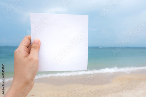 Woman hand holding white paper with sea beach and blue sky background.
