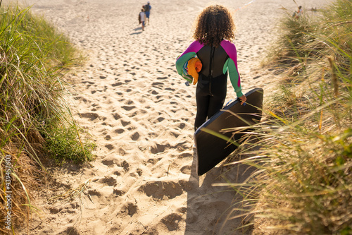 Curly haired girl in wetsuit carries boogie board to beach photo