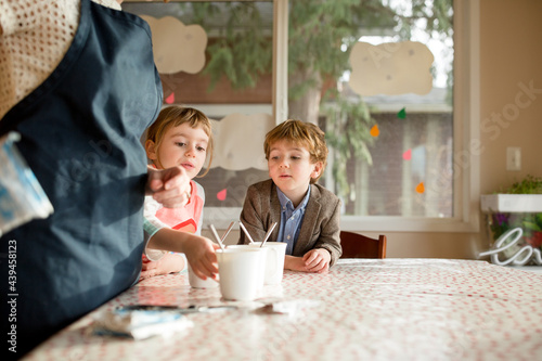 Children look expectantly at mugs on a table while mother stands nearby photo