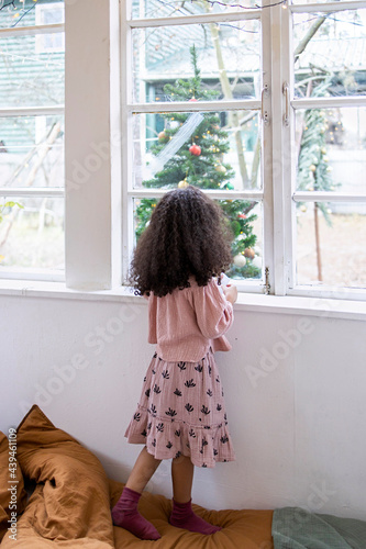 Little afro girl standing in a bedroom and watching outside through the window photo