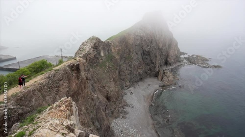 Summer, 2019 - Rudnaya Pristan, Primorsky Region - Panoramic shot of Cape Briner. Tourists walk along a narrow path on the sheer cliff of Cape Briner in the fog. photo