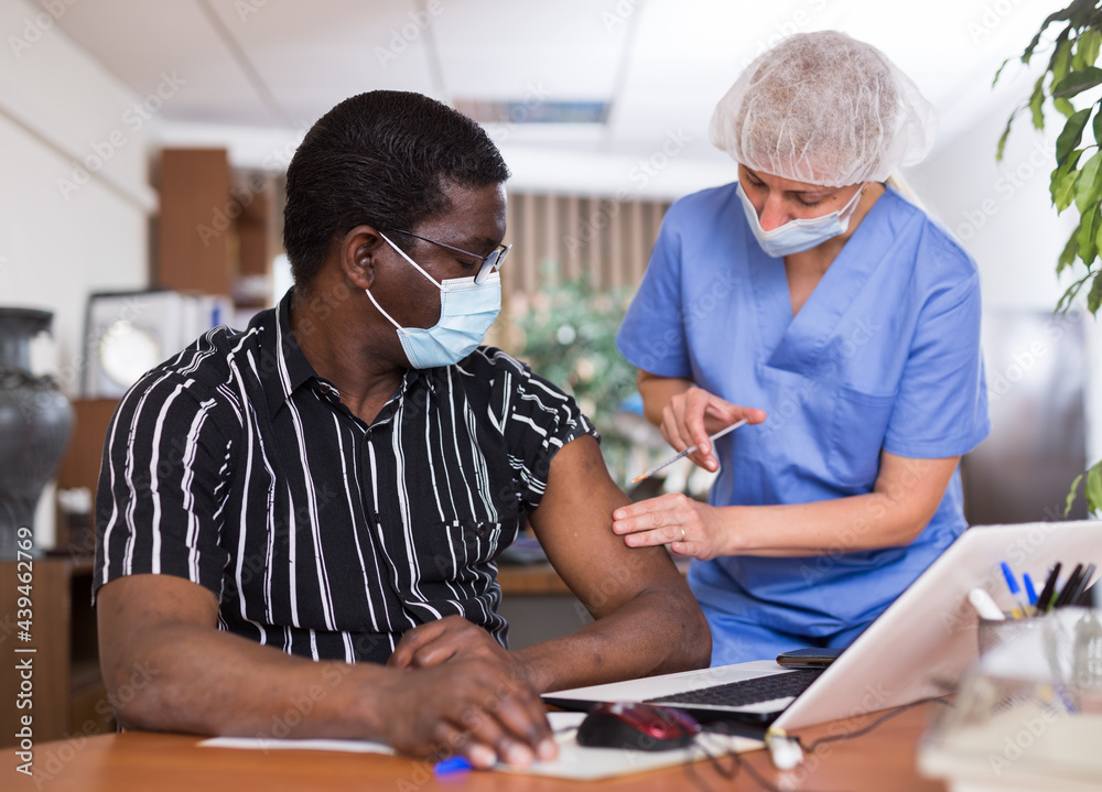 Obraz premium Medical worker who came to the office during the pandemic vaccinates an African male employee in a protective mask ..against the coronavirus