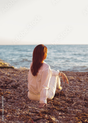 Young smiling woman wearing white shirt sitting on sea shore relaxing and watching sea landscape sunset