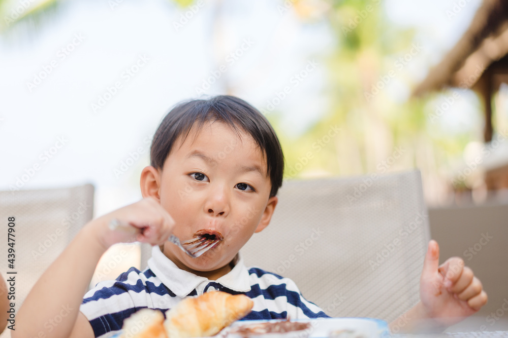 Asian toddler boy eating croissant with chocolate lunch in the school cafeteria.happy asian boy child eating bakery have fork in his mouth.Hungry and Delicious face.Korean japanese boy childhood eat. Asian toddler boy eating croissant with chocolate lunch in the school cafeteria.happy asian boy child eating bakery have fork in his mouth.Hungry and Delicious face.Korean japanese boy childhood eat.