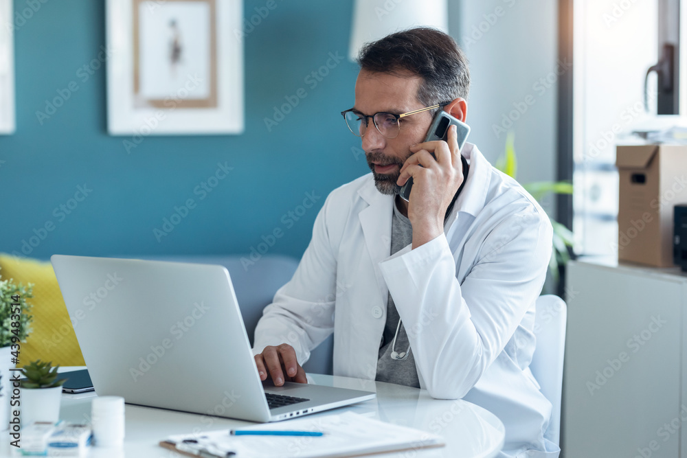 Shot of mature concentrated attractive male doctor working with her computer while talking with smartphone in medical consultation.