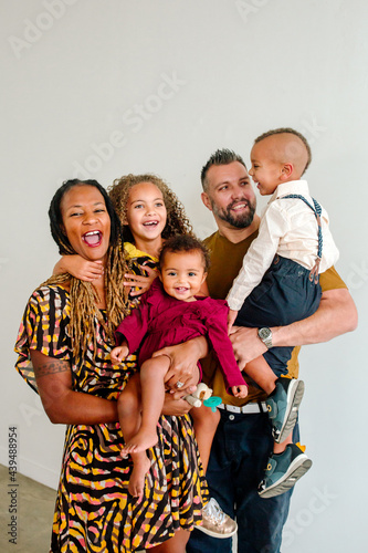 Laughing biracial family with three young children held by dad and mom indoors with neutral background. photo