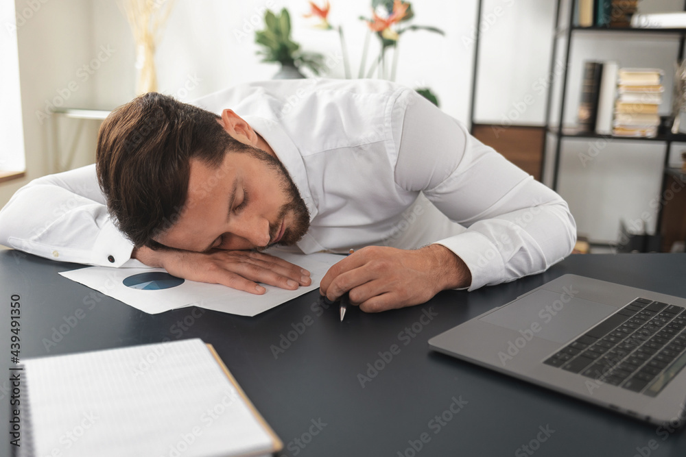 Tired young caucasian businessman fell asleep at the desk in the office analyzing financial report, exhausted man needs sleep Tired young caucasian businessman fell asleep at the desk in the office analyzing financial report, exhausted man needs sleep