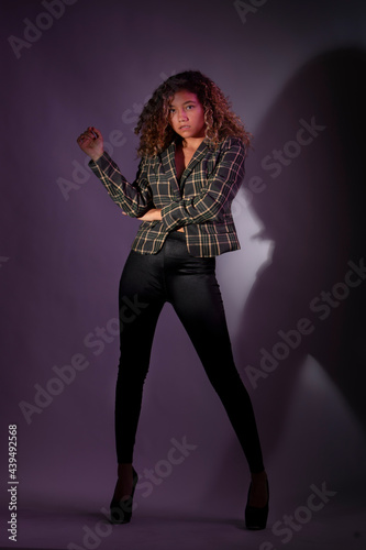 vertical portrait of a curly model wearing a jacket, black pants and high heel shoes in a purple backgroung
