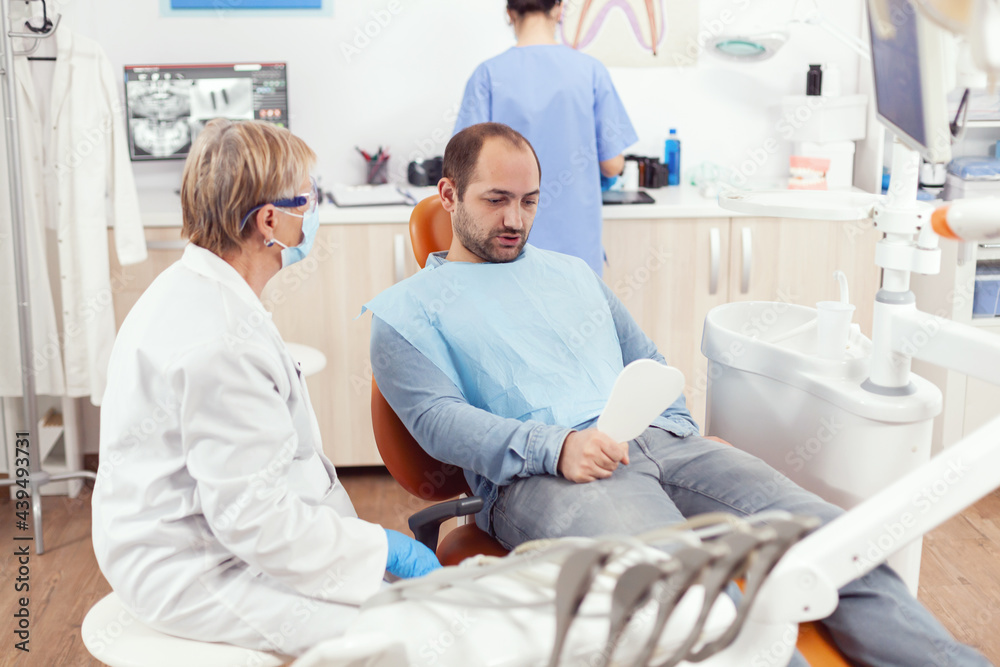 Obraz premium Man patient looking in mirror after medical stomatological team finising dental surgery during stomatology procedure. Senior doctor explaining healthcare treatment for toothache pain