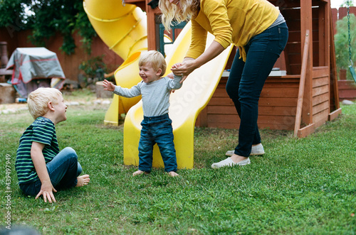 Mom helps happy young son walk to big brother near backyard playset  photo