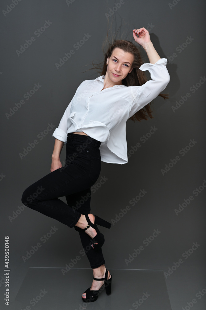 Portrait of a Caucasian girl with fluttering hair in the wind in black trousers and a white blouse with a smile on a gray background in full growth. Studio photo in high resolution. Portrait of a Caucasian girl with fluttering hair in the wind in black trousers and a white blouse with a smile on a gray background in full growth. Studio photo in high resolution.