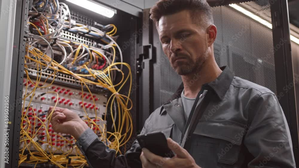 Chest-up of male Caucasian engineer wearing grey worker jacket, looking at smartphone and checking wires in server room