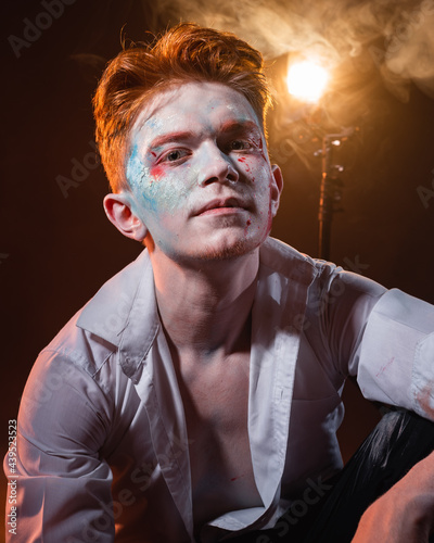 Mad lunatic with a chain on his arm and neck, with makeup on his face against the background of smoke illuminated by yellow light