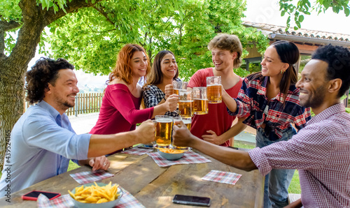 multiethic group of people enjoying beer together outdoor sitting on a garden table. diverse happy friends having fun making a toast drinking alcohol in summer. social, holidays and youth concept