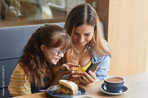 Warm-toned portrait of loving mother and daughter with down syndrome using smartphone while enjoying time together in cafe  copy space