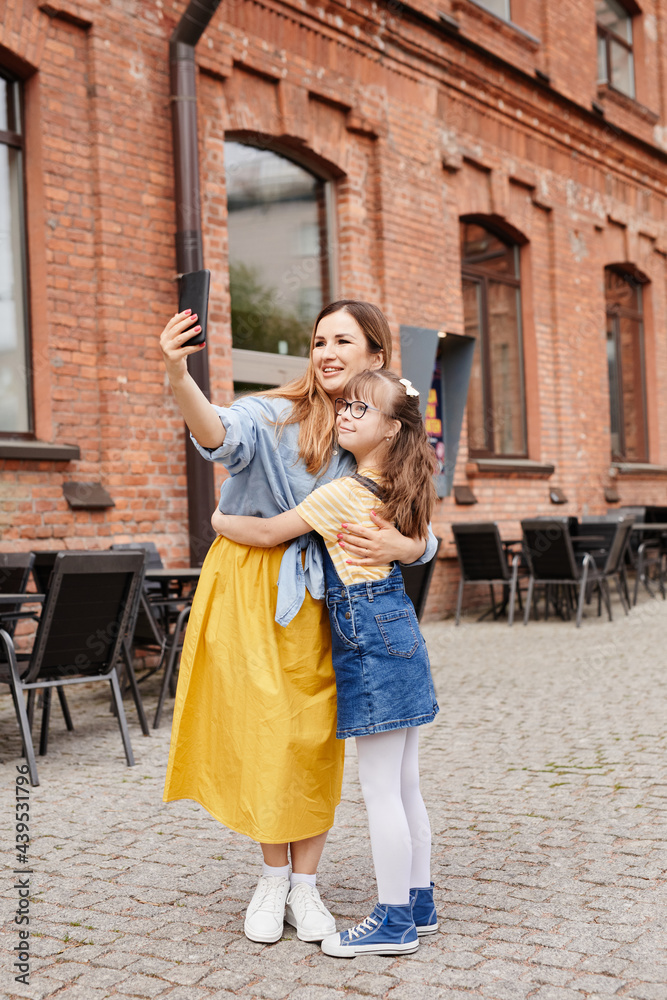 Full length portrait of happy mother and daughter with down syndrome taking selfie together outdoors in city