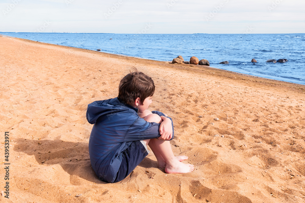 Boy in blue jacket and shorts barefoot sitting on the sand on the seashore, sunny day