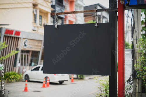 An empty black shop sign hangs on a red iron pole with the blurred background of the street in front of the shop