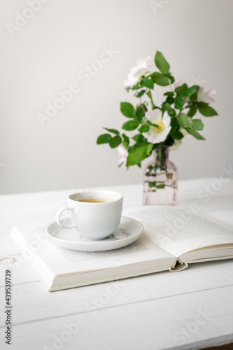 Eine weiße Tasse Kaffee auf einem offenen Buch und frische Blumen in einer Vase. Stillleben, Zuhause.