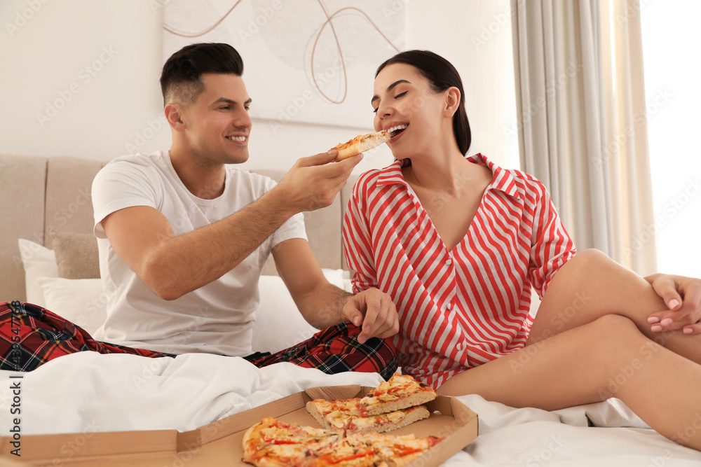 Happy couple in pyjamas eating pizza on bed at home