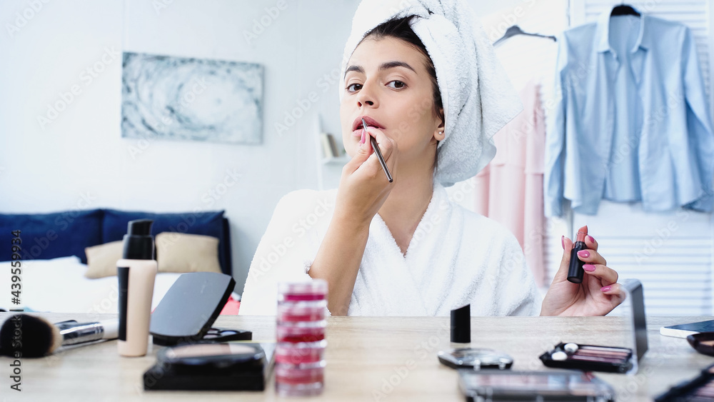 young woman applying lipstick with cosmetic brush near table with decorative cosmetics in bedroom