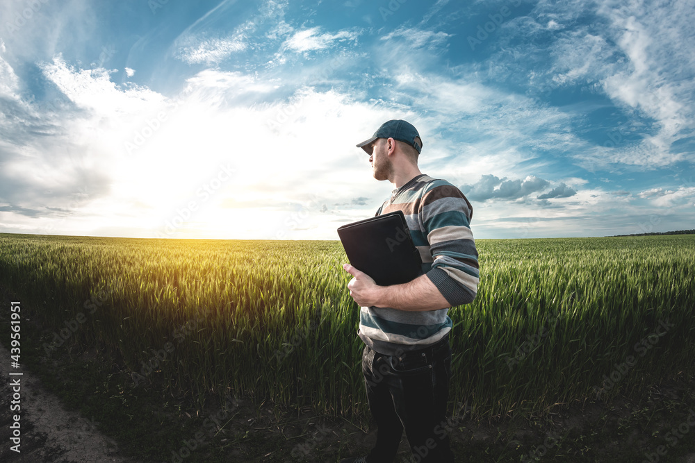 A young agronomist holds a folder in his hands on a green wheat field. A farmer makes notes on the background of agricultural land during sunset. Man in a cap with a folder of documents A young agronomist holds a folder in his hands on a green wheat field. A farmer makes notes on the background of agricultural land during sunset. Man in a cap with a folder of documents
