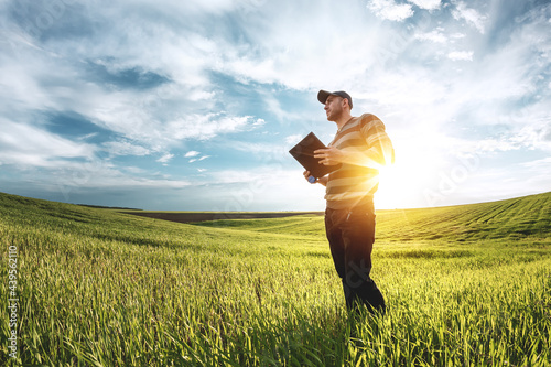 A young agronomist holds a folder in his hands on a green wheat field. A farmer makes notes on the background of agricultural land during sunset. Man in a cap with a folder of documents photo