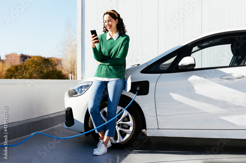 Woman using smartphone near electric car on charging station photo