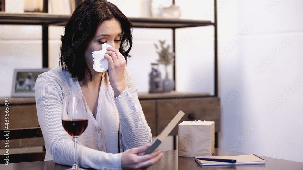 upset woman sitting on table, holding photo frame and wiping tears with paper napkin at home upset woman sitting on table, holding photo frame and wiping tears with paper napkin at home