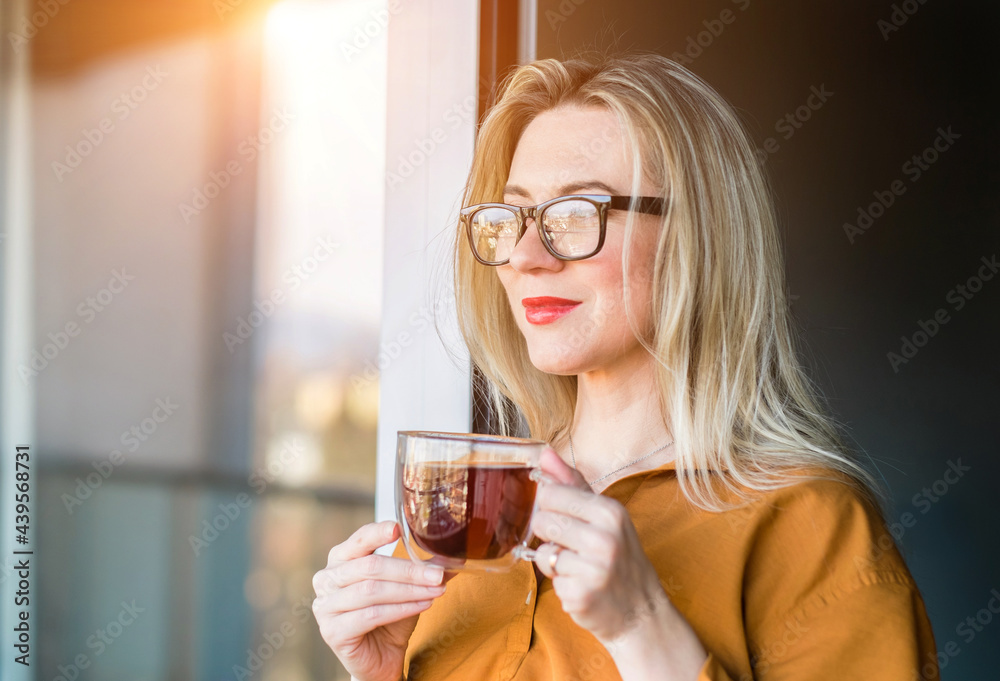 Beautiful smiling Businesswoman in glasses standing near the window and drinking tea cup. Female pretty face, work from home, social distancing, alone at home in office. close up front portrait Beautiful smiling Businesswoman in glasses standing near the window and drinking tea cup. Female pretty face, work from home, social distancing, alone at home in office. close up front portrait