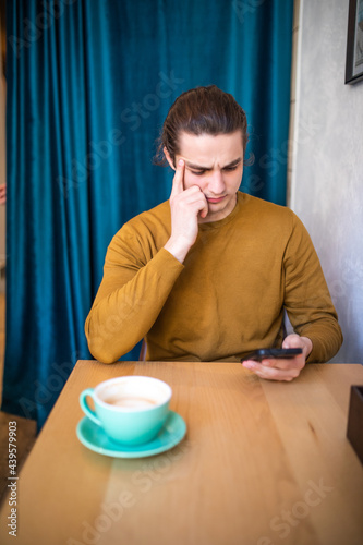 Young thinking man using phone and holding coffee cup