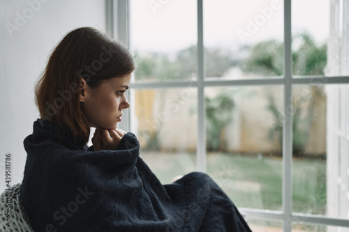 a woman near the window of the house hiding behind a blanket looks out the window