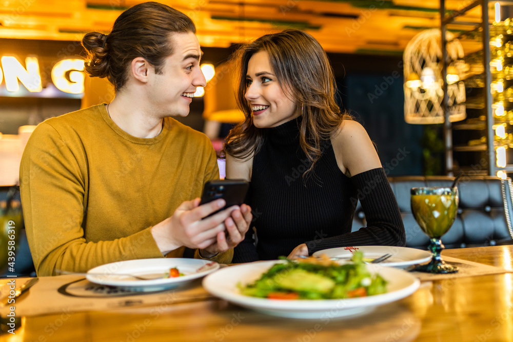 Cheerful young couple browsing internet on smartphone during romantic dinner at cozy cafe. Handsome boyfriend and girlfriend watching video together on cellphone while having lunch Cheerful young couple browsing internet on smartphone during romantic dinner at cozy cafe. Handsome boyfriend and girlfriend watching video together on cellphone while having lunch