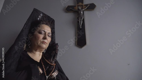 A middle-aged woman is praying with a rosary next to a crucifix.