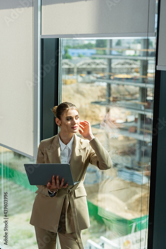 dreamy businesswoman holding laptop while looking away through window in office