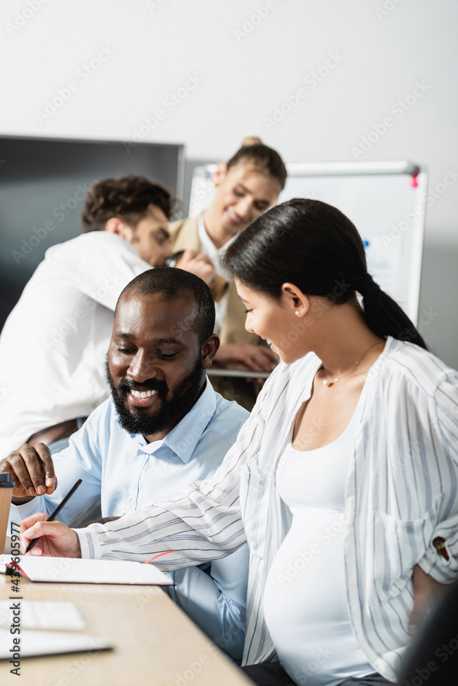 Obraz premium pregnant businesswoman writing in notebook near african american colleague and blurred coworkers