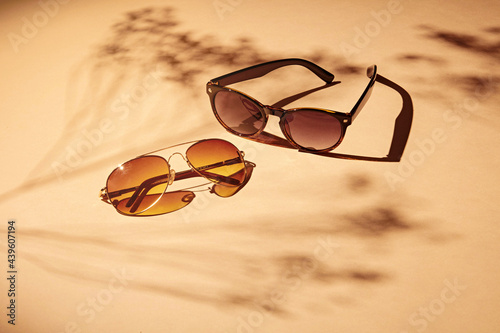 2 pairs of women sunglasses on a soft orange background and strong shadows of flowers