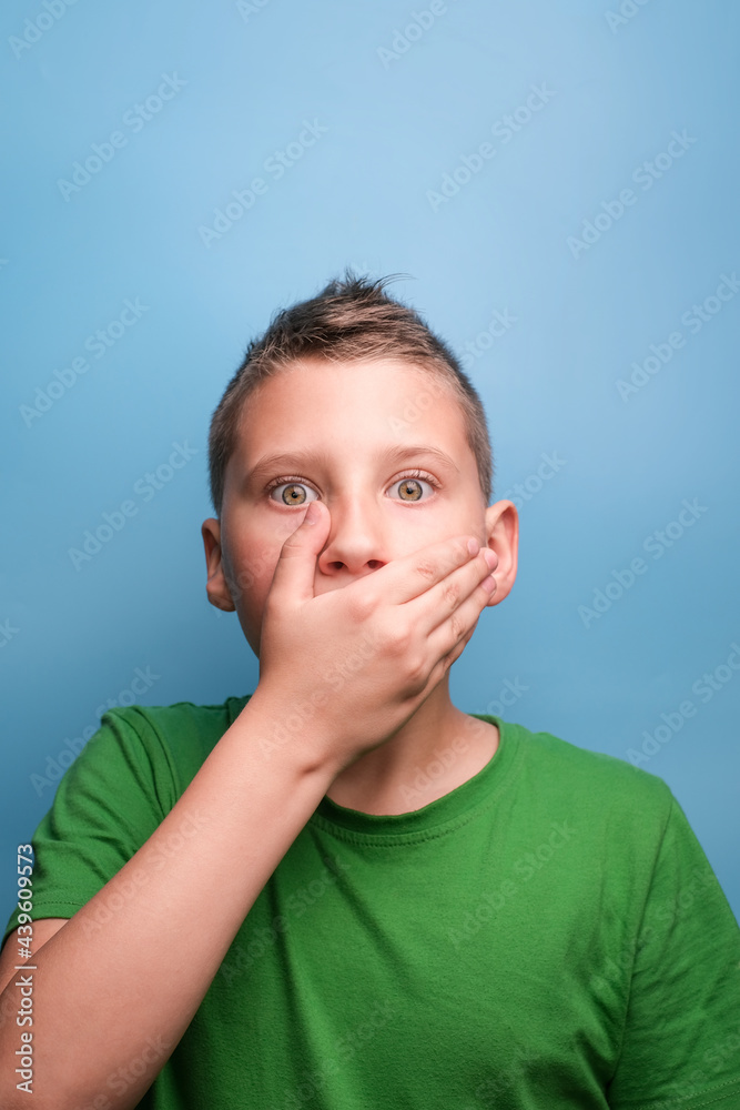Studio shot of emotional adorable boy covering open mouth with hand being surprised and shocked, showing true emotion. Portrait on blue background Studio shot of emotional adorable boy covering open mouth with hand being surprised and shocked, showing true emotion. Portrait on blue background