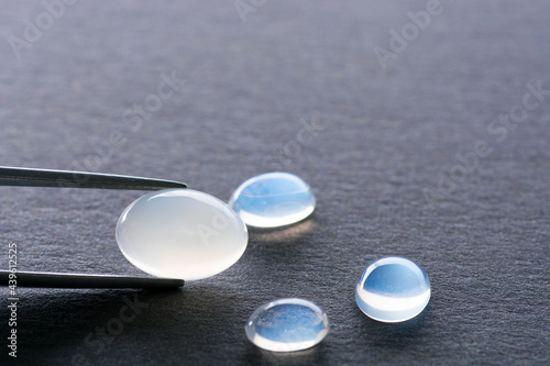 Natural glowing moonstone gems set, lot on gray gradient textured background. Blue adularia oval cabochon loose gesmtones settings and light pink one sample holded in tweezers. Gemology theme. photo