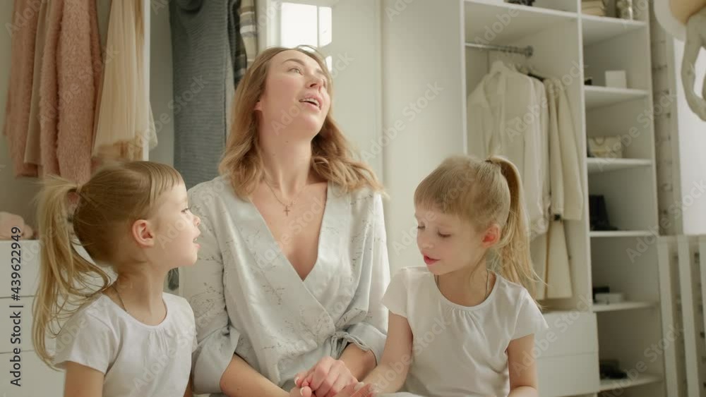 mom and twin daughters in blue pajamas sit on an ottoman against the background of a wardrobe with a lot of clothes