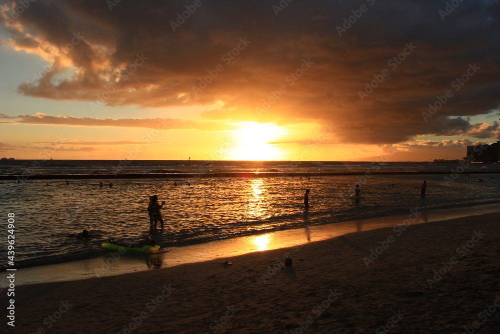 ハワイ　オアフ島。夕暮れのワイキキビーチ。海に沈む夕日と、人々のシルエット。