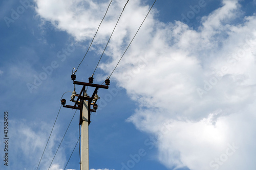 Concrete electric pole with ceramic insulators and voltage lines, in background blue sky with white clouds.