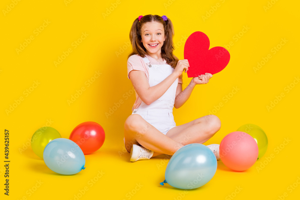 Full length body size photo schoolgirl at party with balloons smiling showing red heart isolated bright yellow color background Full length body size photo schoolgirl at party with balloons smiling showing red heart isolated bright yellow color background