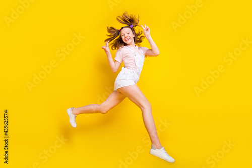 Full length body size view of attractive trendy cheerful girl jumping running showing v-sign isolated over bright yellow color background
