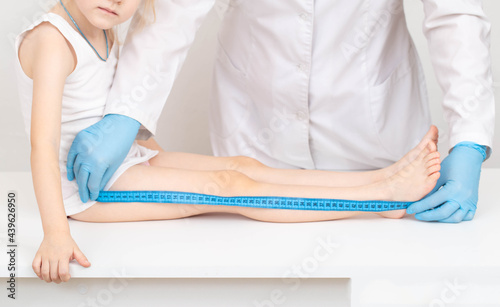 A doctor in blue gloves with a measuring tape measures the length of the legs of a 4-year-old caucasian girl. Child development and growth concept